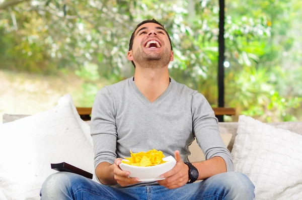 Homem hispânico vestindo camisola azul claro mais jeans jeans sentados no sofá branco segurando tigela de batatas fritas e controle remoto assistindo tv entusiasticamente — Fotografia de Stock