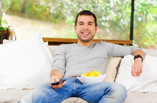 Hispanic male wearing light blue sweater plus denim jeans sitting in white sofa holding bowl of potato chips and remote control watching tv enthusiastically — Zdjęcie stockowe