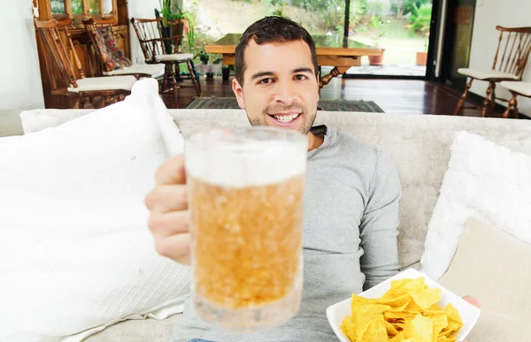 Hispanique heureux mâle portant chandail blye lumière profitant de croustilles de pomme de terre et de bière assis dans le canapé salon blanc face à la caméra — Photo