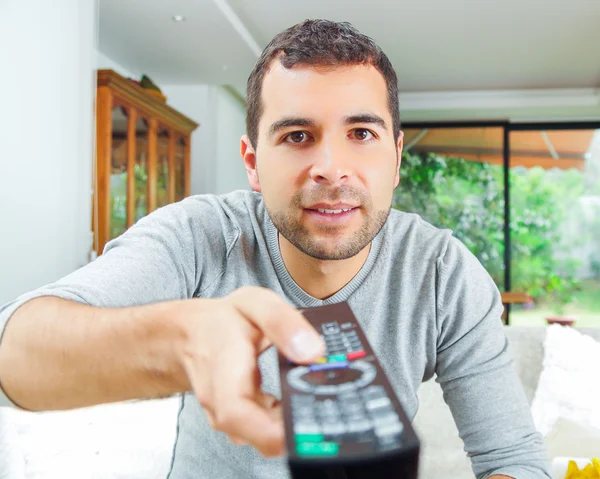 Closeup hispanic male wearing light blue sweater holding up remote control in front of camera, funny angle — Stock Photo, Image