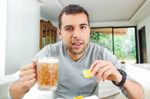 Primer plano masculino hispano usando suéter azul claro sosteniendo cerveza y papas fritas delante de la cámara, ángulo divertido — Foto de Stock