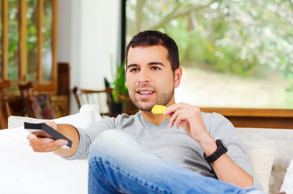 Hispanic male wearing light blue sweater plus denim jeans sitting in white sofa holding potato chip and remote control watching tv enthusiastically — Stockfoto