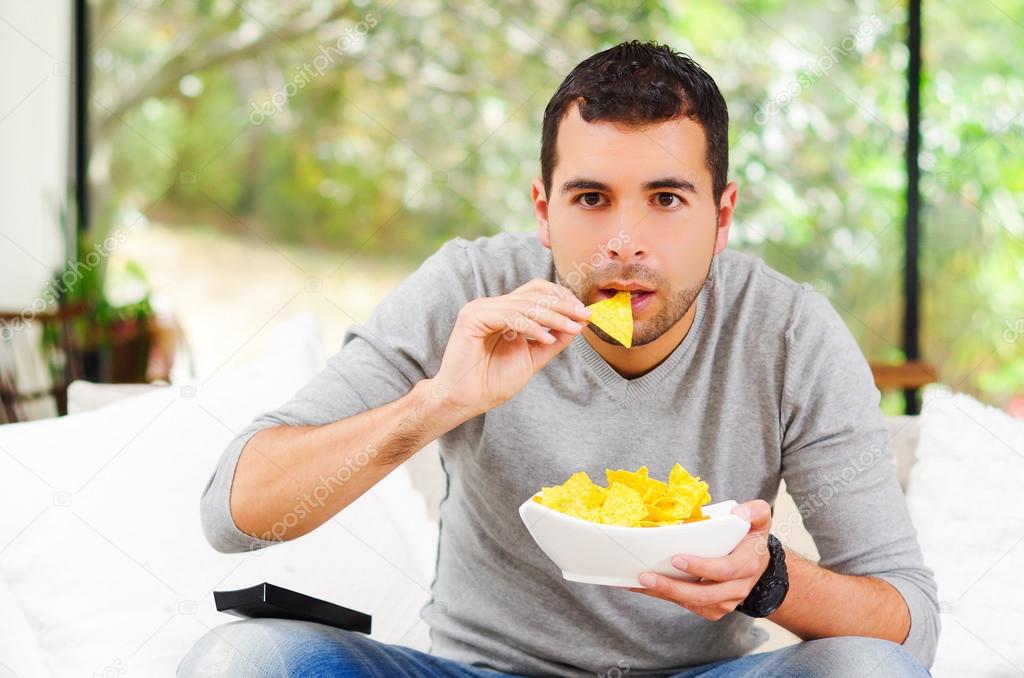 Hispanic male wearing light blue sweater plus denim jeans sitting in white sofa holding bowl of potato chips and remote control watching tv enthusiastically