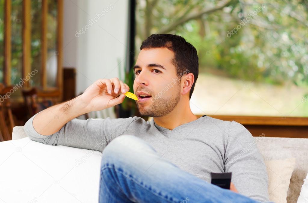 Hispanic male wearing light blue sweater plus denim jeans sitting in white sofa holding potato chip and remote control watching tv enthusiastically