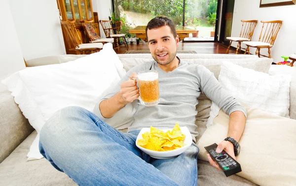 Hispanic pleased male wearing light blye sweater enjoying potato chips and beer sitting in white livingroom sofa facing camera — Stock Photo, Image