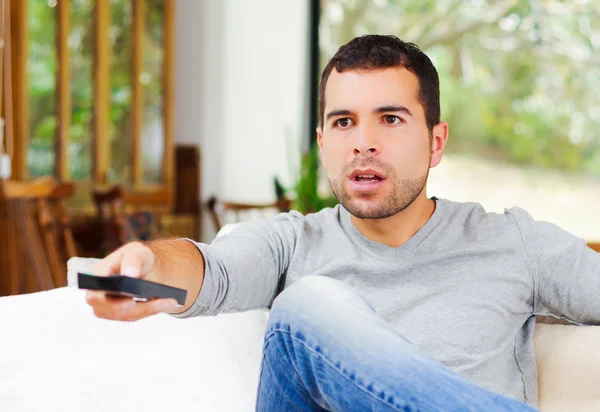 Homem hispânico vestindo camisola azul claro mais jeans jeans sentados no sofá branco segurando batata frita e controle remoto assistindo tv entusiasticamente — Fotografia de Stock
