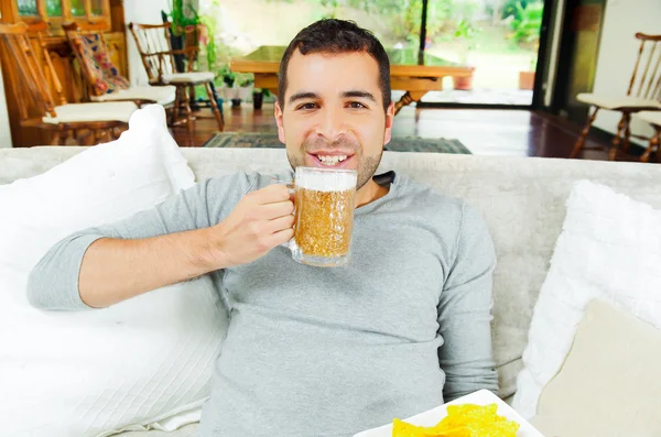 Hispano complacido con un suéter blye ligero disfrutando de papas fritas y cerveza sentado en un sofá blanco frente a la cámara — Foto de Stock