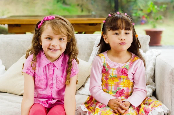 Brunette enfants sœurs assis joyeusement sur canapé blanc salon posant pour appareil photo avec des vêtements typiques rose fille — Photo