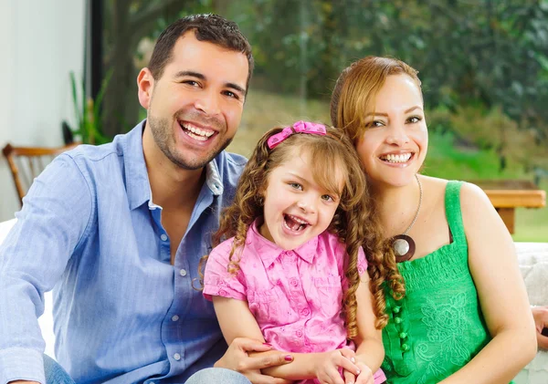 Orgullosos padres hispanos felices posando con una niña linda usando ropa rosa frente al fondo del jardín de la ventana — Foto de Stock