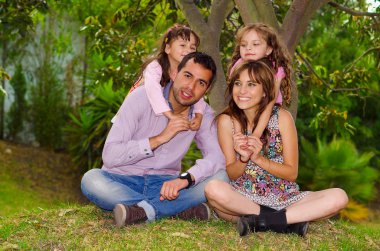 Baba, anne ve iki kızı birlikte Bahçe ortamında, çocuklar onları mutlu bir şekilde gülümseyerek kucaklayan anne arkasında oturan aile portresi