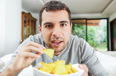 Closeup hispanic male wearing light blue sweater holding up bowl of potato chips in front camera, funny angle
