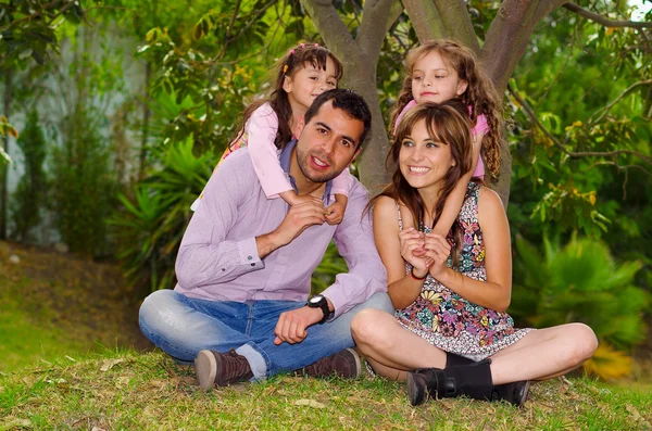 Retrato familiar de pai, mãe e duas filhas sentadas juntas no ambiente do jardim, crianças por trás dos pais abraçando-os sorridentes felizes — Fotografia de Stock