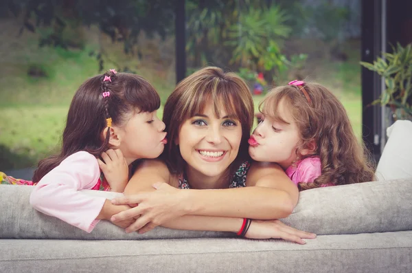 Madre hispana en sofá con dos hijas besándose las mejillas de cada lado, fondo borroso del jardín — Foto de Stock