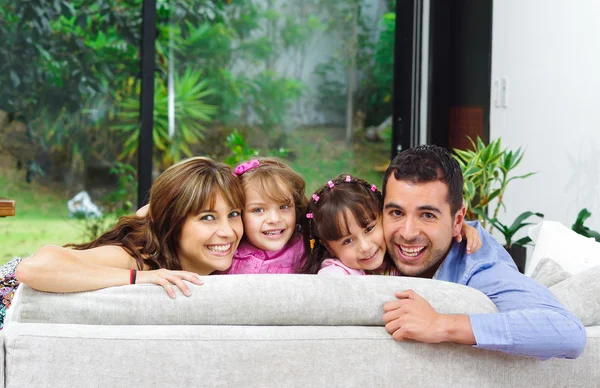 Beautiful hispanic family of four posing with heads sticking up from back sofa looking at camera smiling — Stock Photo, Image