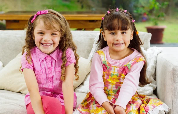 Brunette children sisters sitting happily on white livingroom sofa posing for camera with typical pink girl clothes — Stock Photo, Image