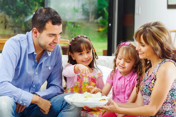 Padres hispanos con dos hijas comiendo de una bandeja de papas fritas sentadas en el sofá mientras sonríen y disfrutan de la compañía —  Fotos de Stock