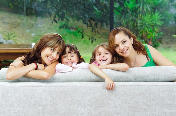 Zwei stolze hispanische Mütter posieren mit ihren Töchtern, die Köpfe ragen vom Sofa hoch und lächeln in die Kamera — Stockfoto