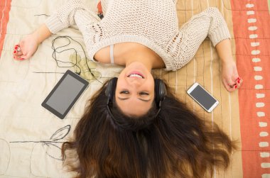 Pretty brunette wearing denim jeans white top lying down on bedsheets daydreaming with smartphone and tablet