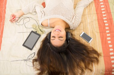 Pretty brunette wearing denim jeans white top lying down on bedsheets daydreaming with smartphone and tablet