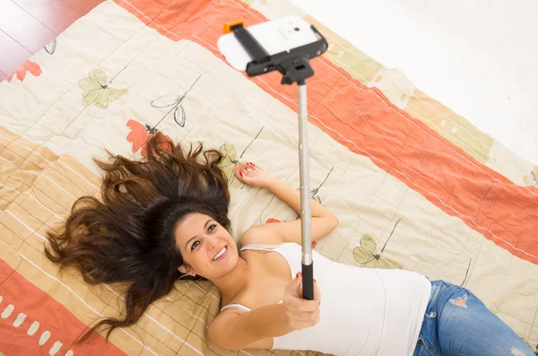 Pretty brunette wearing denim jeans and white top lying down on bedsheets hair spread out, holding selfie stick with mobile phone posing — Stock Photo, Image