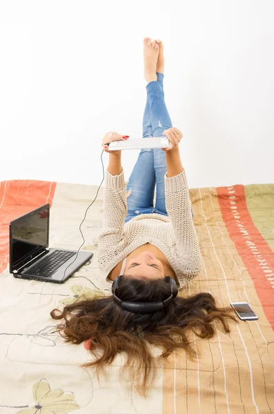 Pretty brunette wearing denim jeans white top lying down on bedsheets daydreaming with smartphone, laptop and tablet — Stok fotoğraf