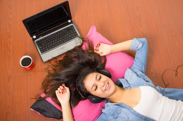Pretty brunette wearing denim clothing and white top lying down wooden surface resting head on pink pillow, laptop plus coffee mug also included — Stok fotoğraf