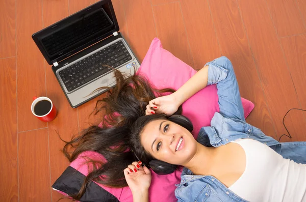 Pretty brunette wearing denim clothing and white top lying down wooden surface resting head on pink pillow, laptop plus coffee mug also included