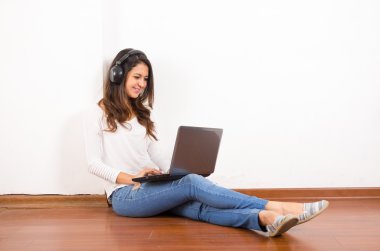 Pretty brunette wearing denim jeans and white top sitting on wooden surface her back against wall, black headphones listening to music using laptop