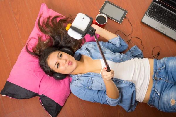 Muito morena vestindo roupas jeans e top branco deitado para baixo superfície de madeira descansando cabeça no travesseiro rosa segurando selfie stick, laptop mais caneca de café também incluído — Fotografia de Stock