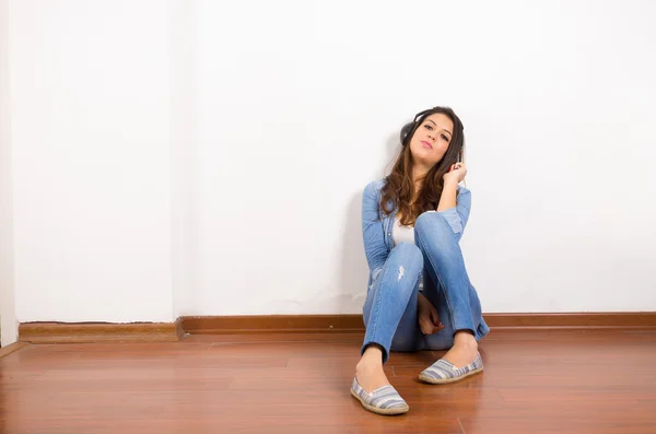 Jolie brune portant un jean et une chemise en denim ainsi qu'un haut blanc assis sur une surface en bois son dos contre un mur, écouteurs noirs écoutant de la musique — Photo