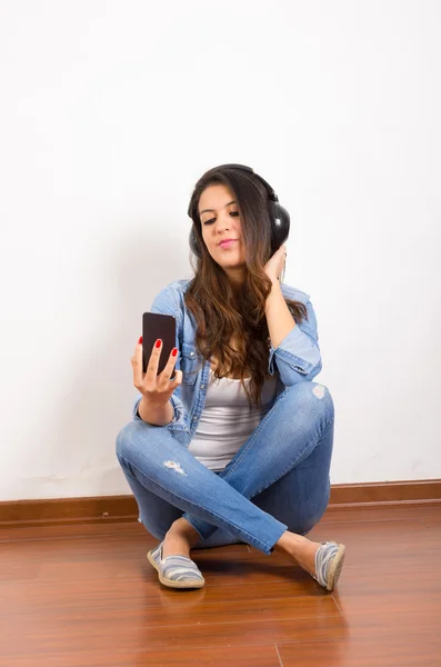 Pretty brunette wearing denim jeans and shirt plus white top sitting on wooden surface her back against wall, black headphones listening to music — ストック写真