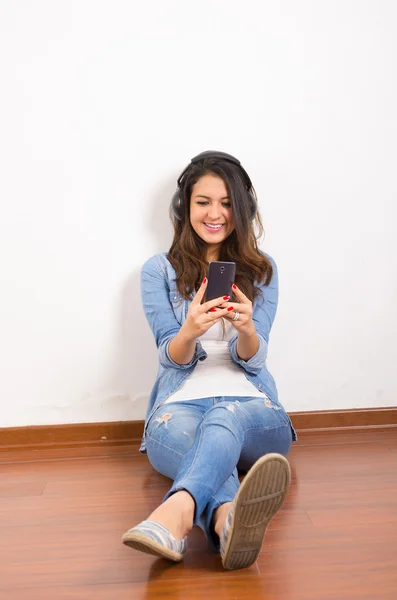 Pretty brunette wearing denim jeans and shirt plus white top sitting on wooden surface her back against wall, black headphones listening to music — Stock Photo, Image