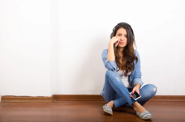 Pretty brunette wearing denim jeans and shirt plus white top sitting on wooden surface her back against wall, black headphones listening to music — Stockfoto