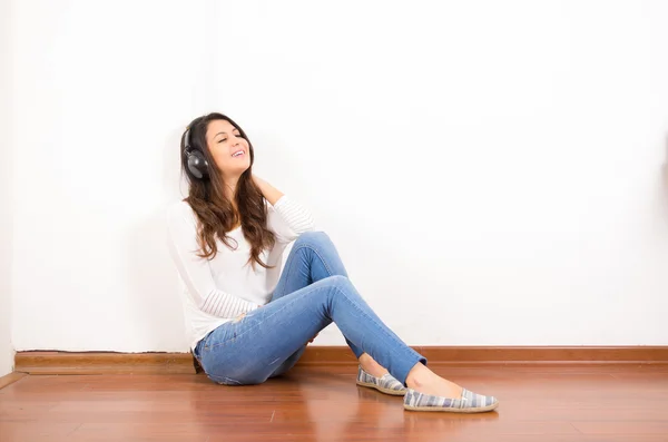 Jolie brune portant un jean denim et un haut blanc assis sur une surface en bois son dos contre un mur, écouteurs noirs écoutant de la musique — Photo