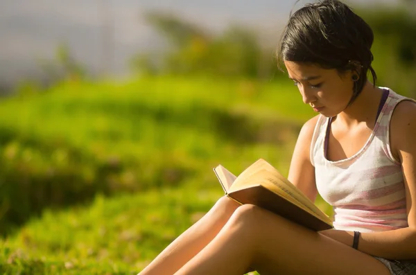 Pretty teenage hispanic girl wearing white top and shorts sitting on grass reading from book — Stok fotoğraf