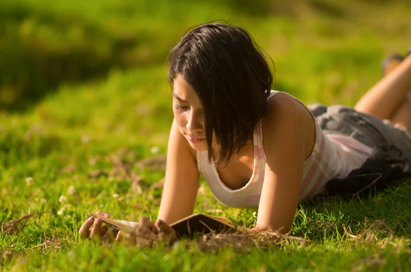 Pretty teenage hispanic girl wearing white top and shorts lying on grass relaxed reading from book — Zdjęcie stockowe