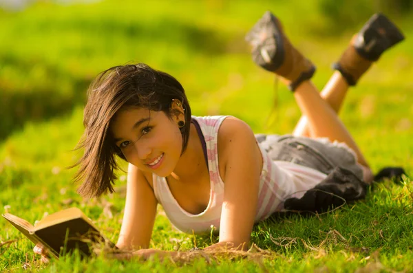 Pretty teenage hispanic girl wearing white top and shorts lying on grass relaxed reading from book — Stok fotoğraf