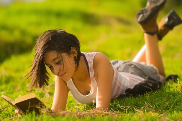 Bastante adolescente hispana chica vistiendo blanco superior y pantalones cortos acostado en la hierba relajado lectura del libro —  Fotos de Stock