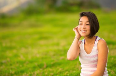 Headshot profile pretty teenage hispanic girl wearing white top with half short hair sitting naturally posing and green park background