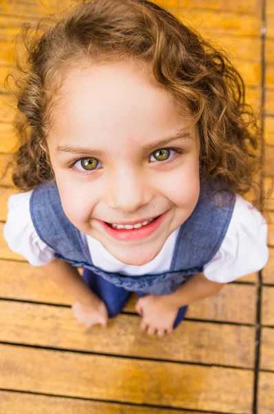 Entzückendes junges brünettes Mädchen, das auf einer Holzoberfläche steht und nach oben in die Kamera blickt, aufgenommen von oben — Stockfoto