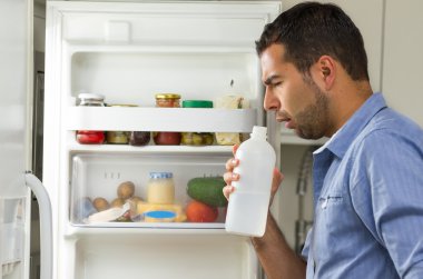 Hispanic male wearing blue shirt standing in fridge door opening smelling a transparent bottle