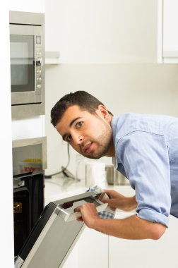 Hispanic male wearing blue shirt in modern kitchen leaning towards open oven door holding a fork looking past camera