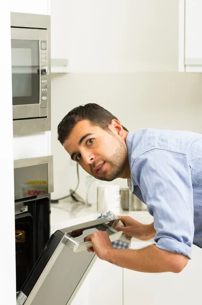 Pria Hispanik mengenakan kemeja biru di dapur modern condong ke arah pintu oven terbuka memegang garpu melihat masa lalu kamera — Stok Foto