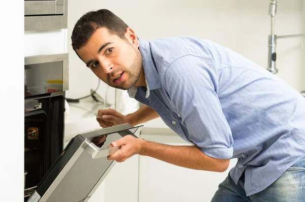 Homme hispanique portant une chemise bleue dans la cuisine moderne penché vers la porte ouverte du four tenant une fourchette regardant derrière la caméra — Photo