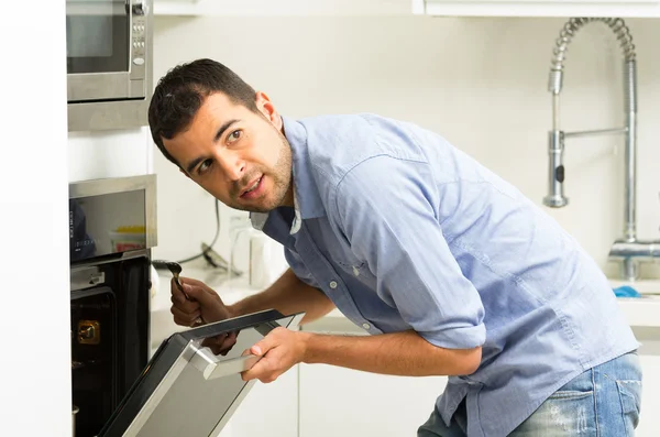 Homem hispânico vestindo camisa azul na cozinha moderna inclinando-se para a porta do forno aberto segurando um garfo olhando para a câmera — Fotografia de Stock