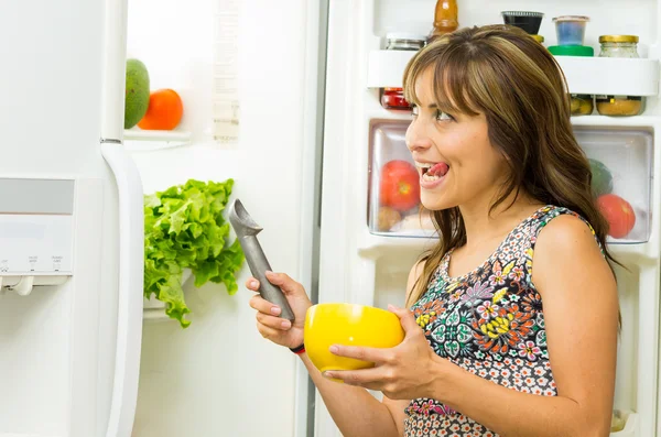 Woman wearing colorful dress in modern kitchen opening fridge door holding an ice cream scoop and yellow bowl — 图库照片