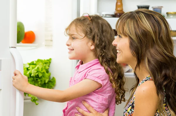 Mother holding up daughter in modern kitchen opening fridge door looking inside happily — Stockfoto