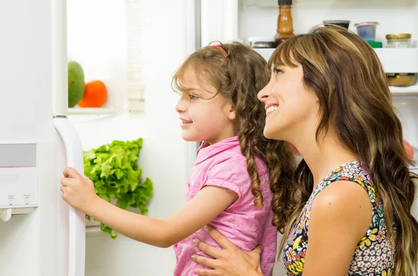 Mother holding up daughter in modern kitchen opening fridge door looking inside happily — Stock Photo, Image