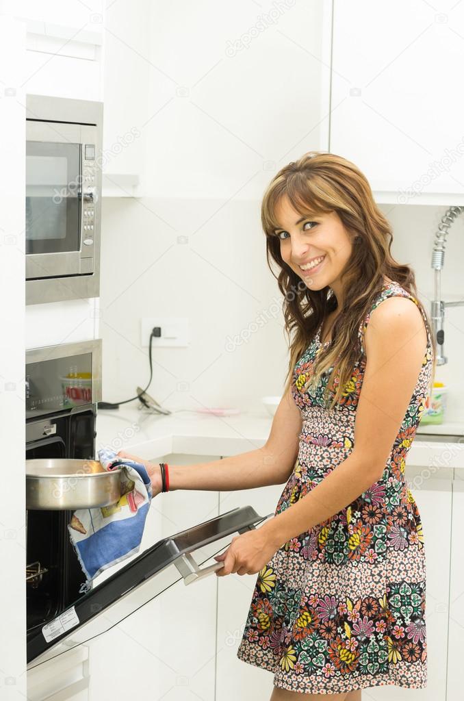 Woman wearing colorful dress in modern kitchen opening oven door holding mittens and cooking pot of metal smiling to camera