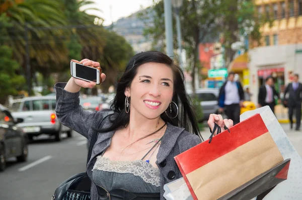 Elegante modelo latina con ropa casual inteligente caminando en la calle urbana sosteniendo bolsas de compras y teléfono móvil sonriendo mientras señaliza para taxi —  Fotos de Stock